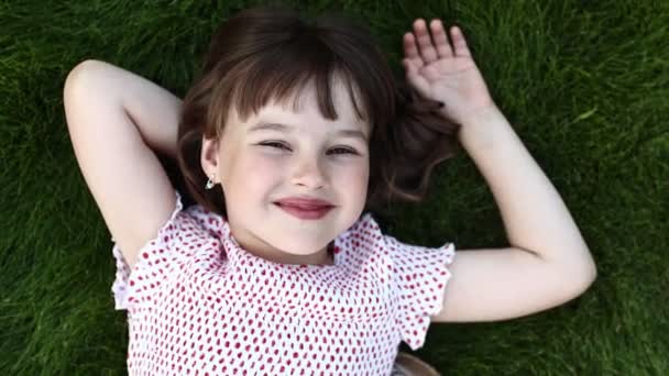 子供の日 夏の暖かい日に緑の芝生の上に寝そべっている幸せなかわいい笑顔の女の子 愛らしい子供は屋外の芝生の上で空を見て夢を見ています 子供の笑顔とカメラを見上げます 幸せな子供時代 — ストック動画