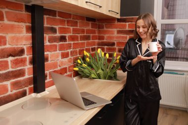 Unrecognized woman is holding and showing finger on white cup of hot drink, coffee or tea with mockup in kitchen at home. Relaxing breakfast time. Bouquet of fresh tulip flowers on background