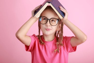 Okul konseptine geri dönelim. Gözlüklü, gülümseyen küçük okul kızı, pembe arka planda izole edilmiş bir çatı gibi kitabı başının üstünde tutuyor. Çocukluk yaşam tarzı konsepti. Okuldaki eğitim. 1 Eylül.