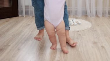 Erkek bebek ilk adımlar için annesinin desteğiyle öğreniyor ve yürüyor. Sağlıklı vücut geliştirme ve kapalı alanda büyüme. Anne, yeni yürümeye başlayan çocuğa denge, bakım ve yürüyüş öğretmek için el ele tutuşuyor..