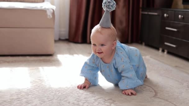 元気いっぱいの小さな笑顔の赤ちゃん青い花のドレスと誕生日コーンの帽子を身に着けている1歳の女の子は ふわふわのカーペットで床を這うポンポン映画で帽子 面白いです赤ん坊子供祝い彼女の最初の休日 — ストック動画