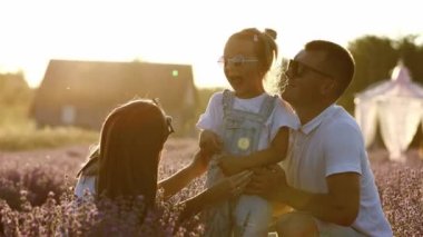 Family day. Happy young mom and dad tickle and hug their little daughter, enjoy relaxing in lavender field at sunset. Parents having fun with kid on summer holiday. Fathers, children, mothers day.