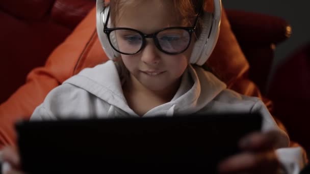 興奮した10代の女の子はソファに座ってデジタルタブレット技術デバイス上で自宅でゲームをプレイします ヘッドフォンやメガネの感情的な子供は インターネットサーフィンパッドコンピュータを保持しています — ストック動画