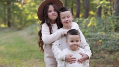 Portrait of happy adorable kids outdoors. Siblings, two stylish little brothers with their older sister hugging together in autumn park, smiling and looking at camera. Happy childhood concept.