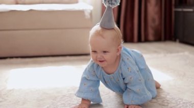 Energetic little smiling baby 1 year old girl dressed in blue floral dress and birthday cone hat with pompon movie crawl by floor with fluffy carpet. Funny babe child celebrates her first holiday.