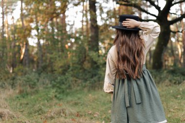 Şık, eski tarz bir elbise giymiş kız çocuğunun arka planı doğa arka planında siyah şapkayla el ele tutuşuyor. Küçük model parktaki tasarımcı elbisesini gösteriyor. Katalog için fotoğraf. Güzellik ve moda konsepti.