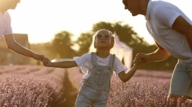 Family day. Happy young mom and dad holding hands and kissing their little daughter, enjoy relaxing in lavender field at sunset. Parents with kid enjoying summer holiday vacation. Children day.