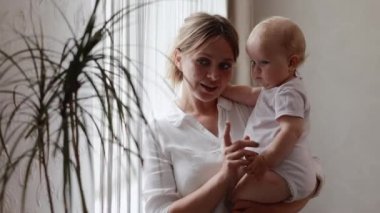 Sevgi dolu genç bir anne kucağında sevimli bir kız bebek kucağında küçük bir çocuğa sarılıyor. Mutlu şefkatli bir anne, evde pencerenin önünde duran küçük çocuğa sarılıyor. Mutlu annelik kavramı.