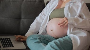 Genç hamile kadın koca göbeğini okşuyor ve dizüstü bilgisayarla evdeki kanepede çalışıyor, internet üzerinden iletişim kuruyor, sosyal medyayı tarıyor. İş, hamilelik ve internet teknolojisi kavramı