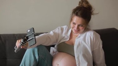 Happy expecting mother and popular vlogger is recording video for online blog sits on sofa in modern flat and showing her big naked belly looks at smartphone camera. Pregnancy and blogging concept.