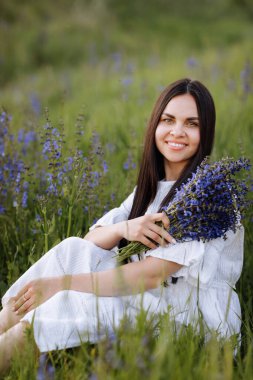 Young smiling beautiful brunette woman in white dress is sitting in green meadow with lupine flowers in spring. Happy girl looks at camera. Women's day. Summertime. Romantic mood. Nature concept.