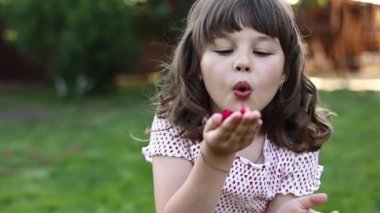 Adorable little child girl 5-6 years old is blowing red roses petals in hands at camera outdoors on summer warm day. Kid having fun at home backyard with green grass. Children day. Happy childhood.