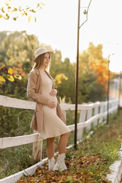 photo of pregnant woman holds hands on belly in the autumn park. Young woman in maternity warm dress and coat waiting for baby birth. Pregnancy, motherhood, mothers day. Happy pregnancy