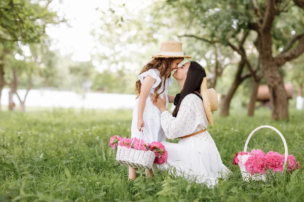 Mothers Day, Womens day. Young beautiful mother is spending time with little daughter in the green summer park. Mom with child are holding baskets of peonies flowers together on nature. Family look.