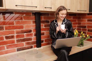 Attractive smiling young woman in pyjama drinking coffee or tea during her breakfast and working with laptop at home. Girl sits on countertop in kitchen. SMM manager enjoying working at home