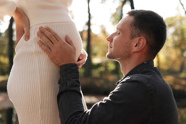 Cropped shot of happy young husband enjoying strokes, talks and kisses tummy of wife, waiting for birth baby outdoors in sunbeams in the city park. Maternity prenatal care and woman pregnancy concept