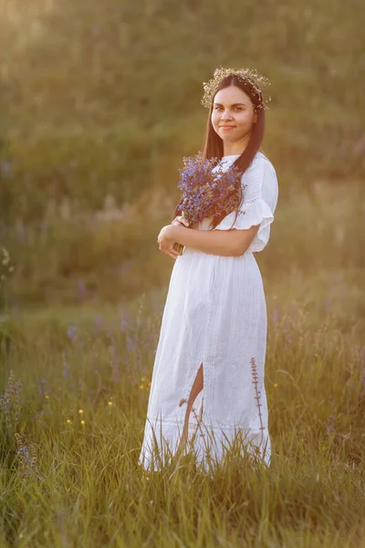 Young smiling beautiful woman in white dress and wreath of wildflowers with a bouquet of purple lupine flowers stands in green meadow in spring. Womens day. Summertime. Romantic mood. Nature concept.