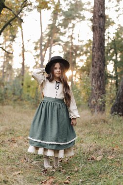 Portrait of little caucasian girl looking at camera standing outdoors. Stylish adorable smiling child in black hat enjoying childhood testimonial concept at green park background