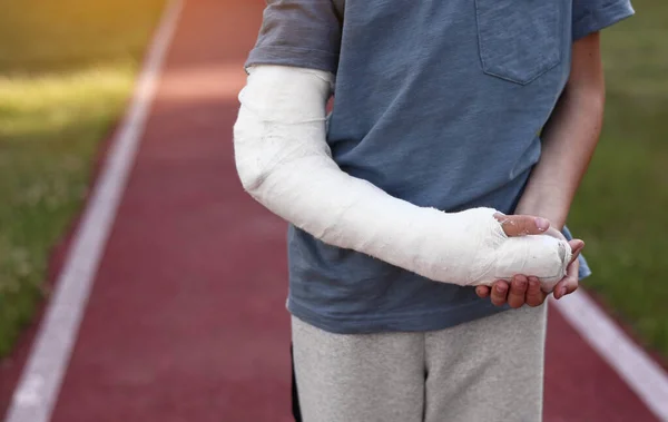 cropped photo of little boy with broken hand outdoors on sports ground on summer day. 9 years child with a fractured limb. plaster cast on the arm. health and medical concept.