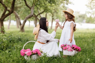 Mothers Day, Womens day. Young beautiful mother is spending time with little daughter in the green summer park. Mom with child are holding baskets of peonies flowers together on nature. Family look.