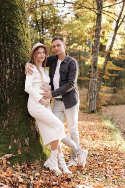 Gülümseyen şık, hamile bir kadın ve erkek kucaklaşır, karnına dokunur ve bebeğin tekmelerini dinler, sonbahar parkında dışarı çıkarlar. Hamilelik, annelik ve ebeveynlik kavramı. Aile doğmamış bir bebek bekliyor.