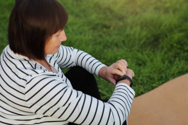 World health day. Mature adult woman on yoga mat looking at her smart watch to check exercise progress after morning routine outdoors on nature. Senior healthy lifestyle, vitality, healthcare concept.