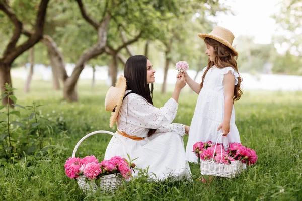 Mothers Day, Womens day. Young beautiful mother is spending time with little daughter in the green summer park. Mom with child are holding baskets of peonies flowers together on nature. Family look.