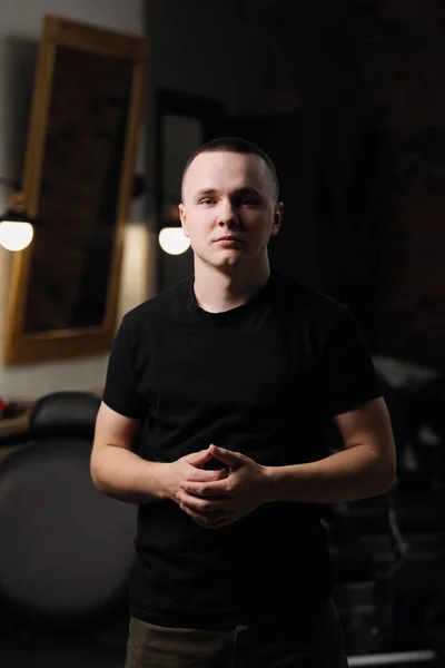 Serious young barber man in black t shirt stands with crossed hands by the work place at barber shop and confidently looking at camera. Owner of small business, hair beauty salon