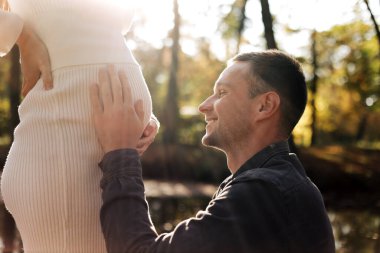 Cropped shot of happy young husband enjoying strokes, talks and kisses tummy of wife, waiting for birth baby outdoors in sunbeams in the city park. Maternity prenatal care and woman pregnancy concept
