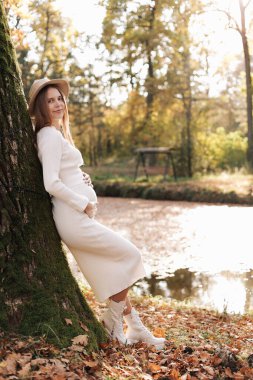 Elegant young pregnant woman in white knit dress and hat touching and stroking belly, relaxing in park by the lake on autumn warm day. Preparation for childbirth, healthy motherhood. Mothers day.