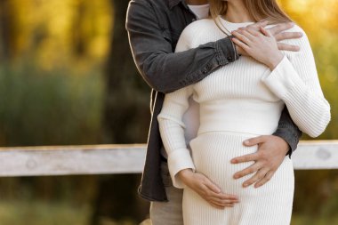 Mutlu stil sahibi hamile kadın ve erkek kucaklaşır, öpüşür, karnına dokunur ve bebeğin tekmelerini dinler, sonbahar parkında dışarı çıkarlar. Hamilelik, annelik ve ebeveynlik kavramı. Aile doğmamış bir bebek bekliyor.