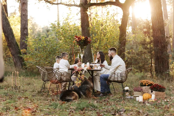 Happy family day. Young dad, mom, child daughter, two little sons, big dog have picnic and sitting by the table in autumn park. Children day. Concept of family coziness, relationships, communication.