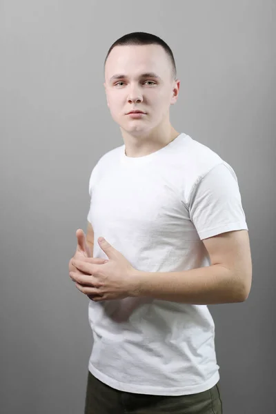 スタジオポートレートのハンサムな自信のある若い男でカジュアルホワイトTシャツでモックアップと彼の手を渡り グレーの背景に隔離されたカメラを見て — ストック写真