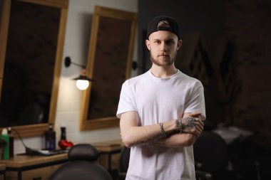 Dövmeli yakışıklı genç berberin portresi berber dükkanında kollarını kavuşturup kameraya bakıyor. Küçük işletme sahibi, kuaför salonu