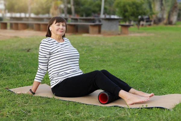 Relaxation after workout. Happy adult sportive woman in casual sportswear works out the fascia of the back muscles of the legs while sitting on a foam massage cylinder outdoors. World health day.