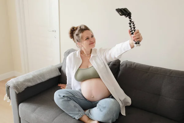 在现代公寓的沙发上 妈妈和流行的维基百科正在为在线博客录制视频 并在智能手机相机上展示她赤身裸体的大肚子 怀孕与博客概念 — 图库照片