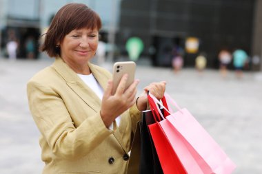 Alışveriş çantaları ve akıllı telefonları olan şık yetişkin kadın blogcu alışveriş merkezi yakınlarındaki sosyal ağlarda takipçilerle iletişim kuruyor. Tüketim, satış, zengin hayat, sanal hayat kavramı. Kara Cuma.