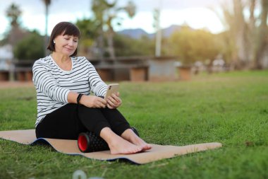 World health day. Smiling adult woman in casual sportswear sitting on yoga mat, using cellphone and relaxing to listen music with headphones after training with massage roller on green grass outdoors.