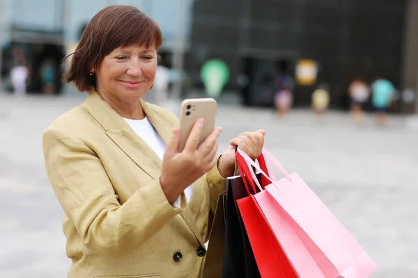 Stock image Stylish adult woman blogger with shopping bags and smartphone is communicating with followers in social networks near shopping mall. concept of consumerism, sale, rich life, virtual life.Black Friday.