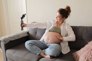 Happy expecting mother and popular vlogger is recording video for online blog sits on sofa in modern flat and showing her big naked belly looks at smartphone camera. Pregnancy and blogging concept.