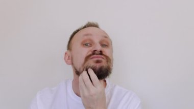 Kesilmemiş genç adam tıraş olmamış çenesini kaşıyarak kameraya bakıyor. Beyaz tişörtlü, kalın sakallı ve bıyıklı, iri yarı, çirkin bir erkek. Güzellik salonu konsepti, berber dükkanı..