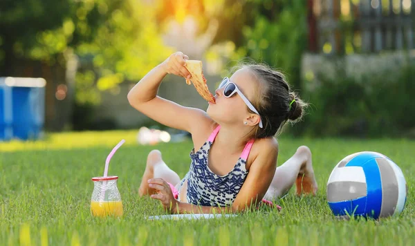 夏天阳光明媚的日子 小女孩躺在草地上吃披萨 在笔记本电脑上玩耍 这孩子在游戏结束后吃午饭 后背吊床及球 — 图库照片
