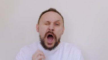 Uykucu, tıraşsız, kılsız, esneyen ve sakalını kaşıyan adam. Sıkılmış erkek, ilgisiz ve ilgisiz bir şekilde beyaz tişörtle beyaz duvardaki kameraya bakarak uyumak istiyor. Sabah partisinden sonra