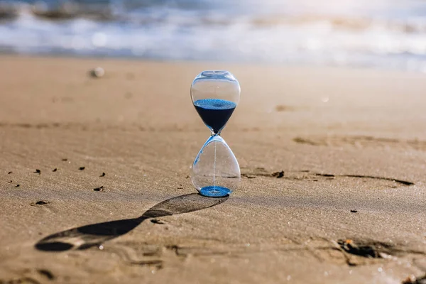 Hourglass with blue sand inside in a warm golden morning sunlight with shadow on a sandy beach and sea background, starting time for a new day or running of time. Concept of the rapid passage of time.
