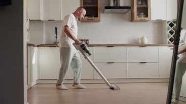 Neşeli genç adam, kablosuz elektrik süpürgesiyle mutfakta yerleri temizlerken müzik dinliyor ve dans ediyor. Temizlik dairesi, ev ve temizlik, süpürge, evi temizle.