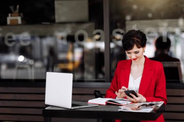 Business young woman in red blazer working on laptop and making notes on cafe terrace. Urban people with modern technology. Successful business. Smart work concept, possibility to relocate activities.