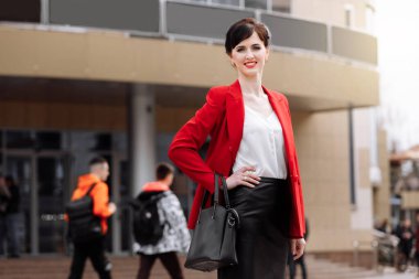 Smiling business woman dressed red jacket, black skirt with leather business bag standing outdoor near corporate office building, hand on the hip. Confident caucasian female business on city street