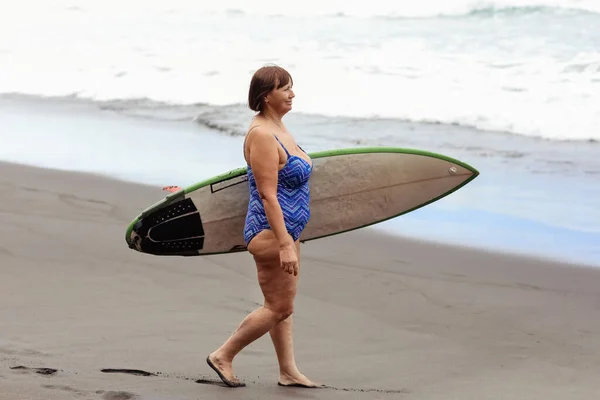 身穿泳衣的成年女子持冲浪板去冲浪 在海边的黑色沙滩享受暑假 中年妇女有积极的生活方式 极端的水上运动课 游泳活动 — 图库照片