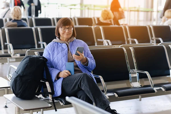 機内持ち込み手荷物 バックパック パスポートをお持ちの大人の旅行者の女性は 出発のためのフライト 空港ターミナルで無料のWi Fiを備えたスマートフォンでのフライト出発時間のオンラインチェックを待っています — ストック写真