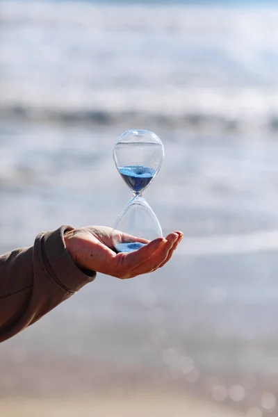 Time is running fast. Hourglass with blue sand inside in mature woman\'s hand symbolize the brevity of life. Background is sea with beautiful sunlights. Concept of the rapid passage of time.Copy space.
